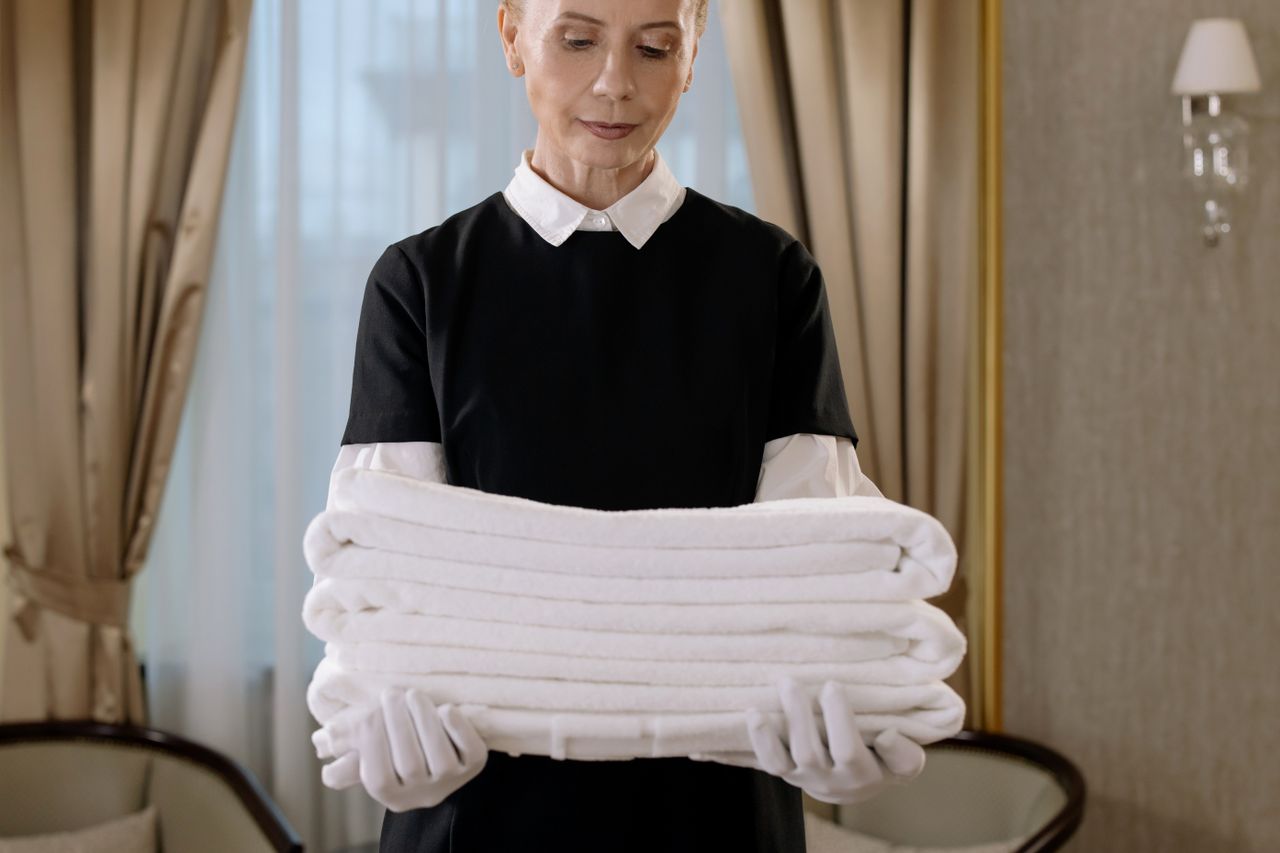 gallery-03 Professional housekeeper holding a stack of fresh towels in a luxurious hotel room, showcasing elegance and service.