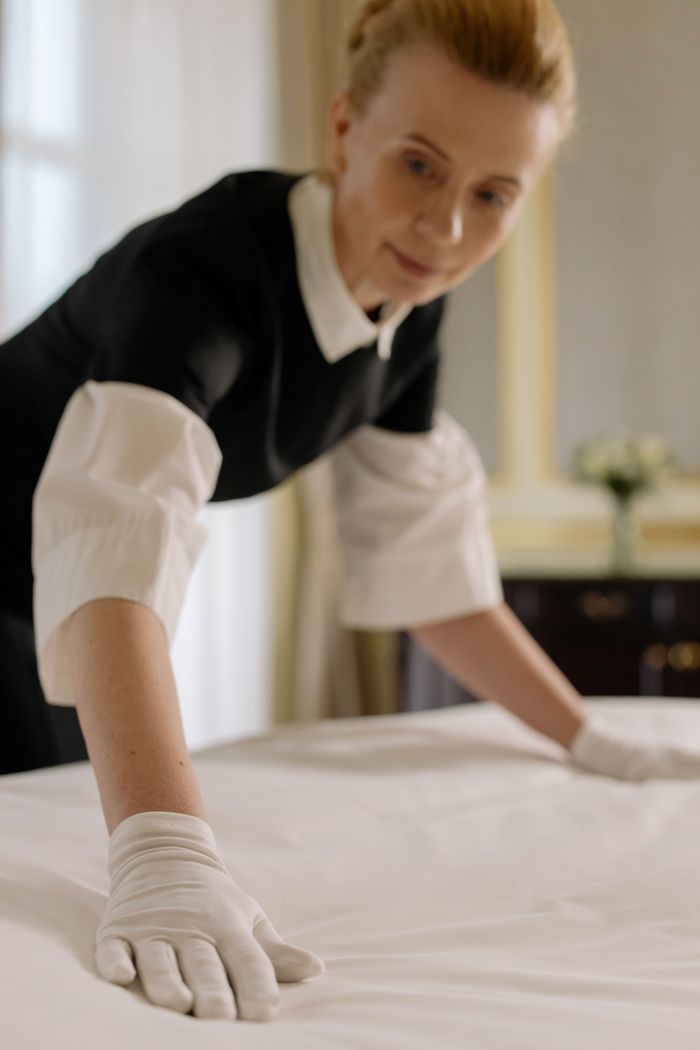 gallery-04 A housekeeper elegantly arranges bed sheets in a luxurious hotel room, showcasing hospitality expertise.